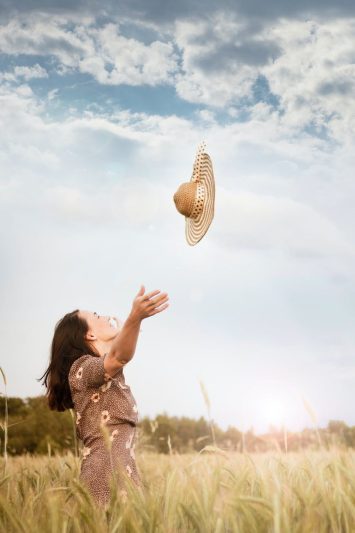 woman standing on wheat field throwing brown sun hat on the air under white and blue skies