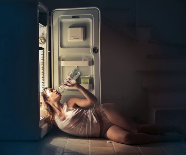photo of woman in white vest and panties lying on floor next to open fridge while drinking water from plastic bottle
