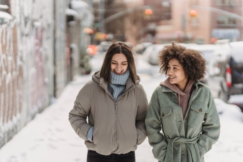 delighted diverse women walking on snowy sidewalk