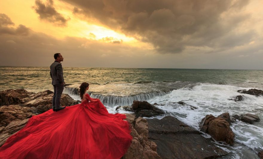 woman in red gown and man in gray suit standing and sitting on boulders in front of ocean