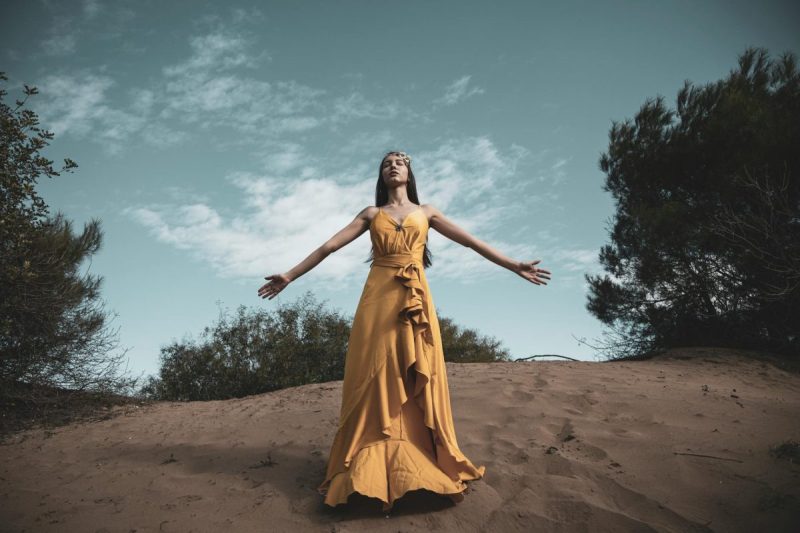 photo of woman in yellow dress standing on brown sand under blue sky with her eyes closed and her arms spread