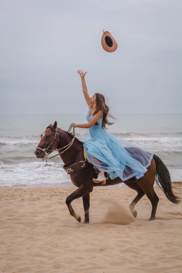 a woman in a blue dress horseback riding on a beach and throwing her hat in the air