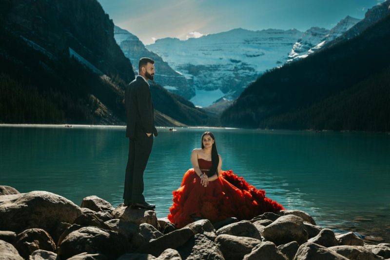 man in a suit and woman in a red dress posing by the lake in mountains