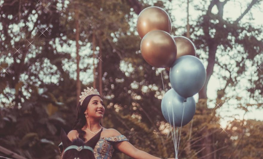 woman in a blue gown and a tiara on her head riding a horse with balloons in her hand