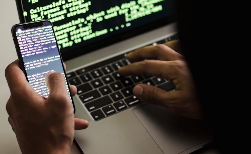 unrecognizable hacker with smartphone typing on laptop at desk