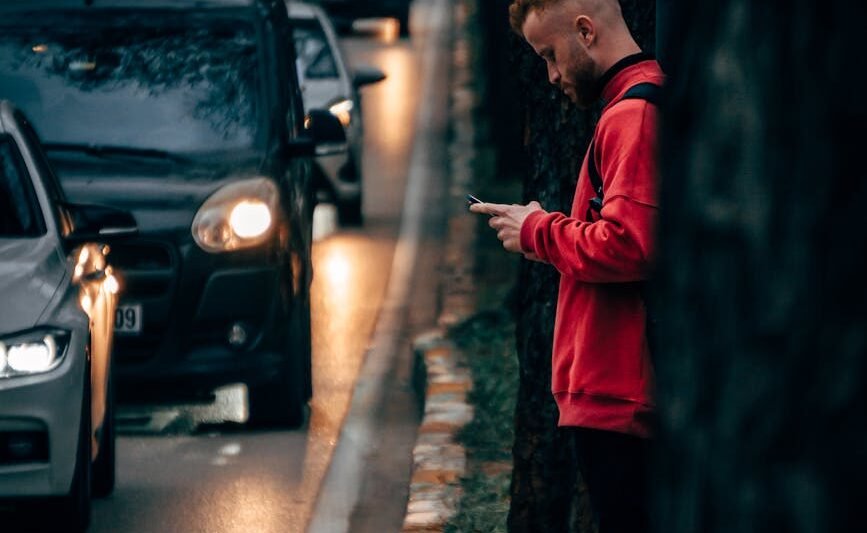 man in red sweater standing near road full of cars while using a phone